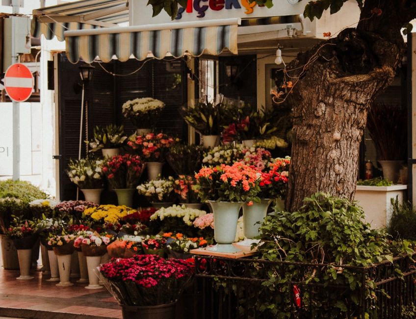 A street-facing flower shop with a display of vibrant, freshly cut floral arrangements in various sizes and styles. The flowers include pink, white, red, yellow, and purple blooms such as chrysanthemu