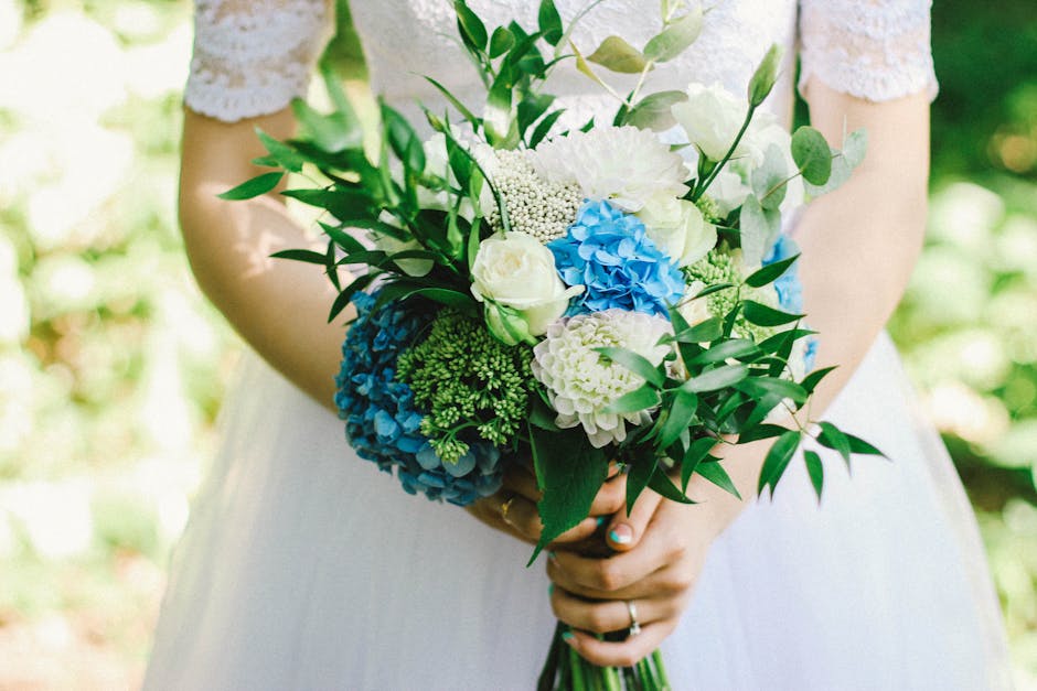 A person in a white dress, likely a bride, holding a bouquet of fresh flowers, including white roses, white hydrangeas, blue hydrangeas, and green foliage. The bouquet is arranged in a rounded, natura