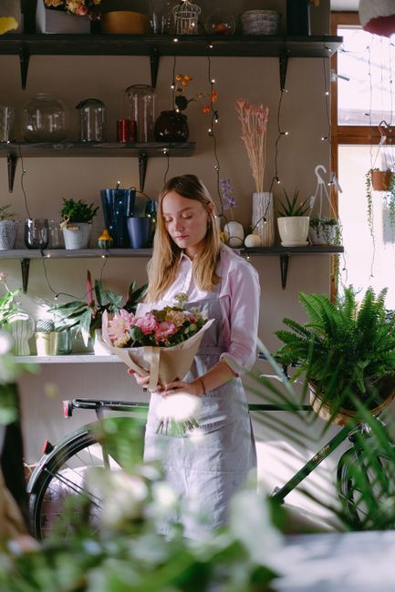 A young woman with light brown hair, dressed in a white blouse and apron, holds a wrapped bouquet of fresh flowers consisting of pink roses, white lilies, and greenery. She stands indoors in a flower 