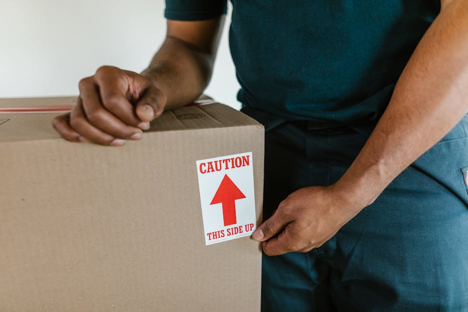 Close-up of a person's hands handling a cardboard box with a caution label that has a red arrow pointing upward and the text 'CAUTION THIS SIDE UP.' The individual is wearing a dark green shirt and bl